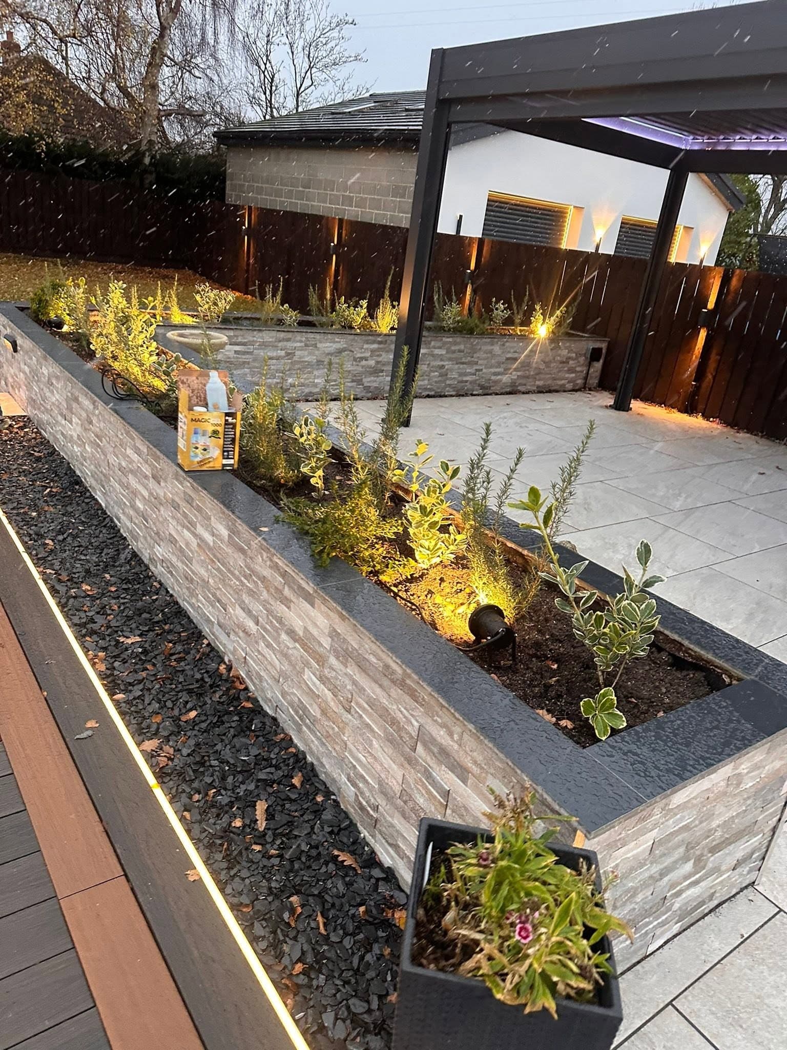 Stone-clad raised planter lit by spike spotlights, with deck-edge LED strip and a louvred pergola behind